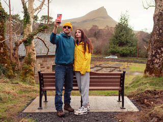 Father and teenager daughter by a bench on a trip taking picture on smartphone. Nature scenery with...