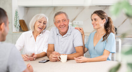 Smiling elderly parents and adult children sit at the table with tea and talk about home life