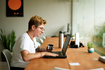 Transgender freelancer works on laptop in modern office, sipping tea from insulated mug. Inked pro...