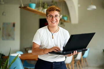 Cheerful transgender professional holds laptop, smiles in modern office. Gen Z using tech for work,...