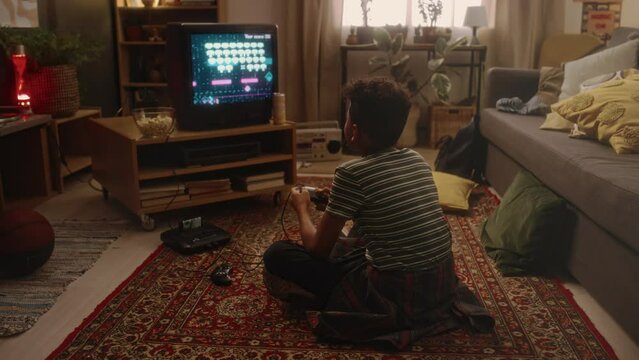 Back full footage of boy sitting on floor and passionately playing space shooter video game on tube TV on retro console at home alone, atmosphere of nostalgic childhood in 90s