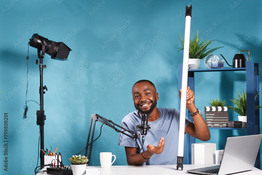 Poster smiling blogger holding tube light equipment and explaining features while recording video for onlin