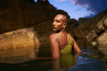 Trans sexual black fashion model poses chest deep inside natural pool surrouded by rocks on tropical island at night. Androgynous ethnic fashion model in the middle of backwater exotic scenic location