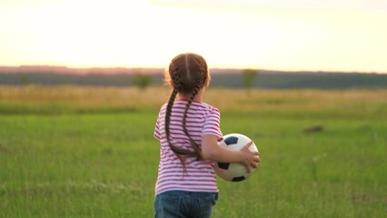 Active cute little girl kid running on sunset grass field with football ball back view closeup....