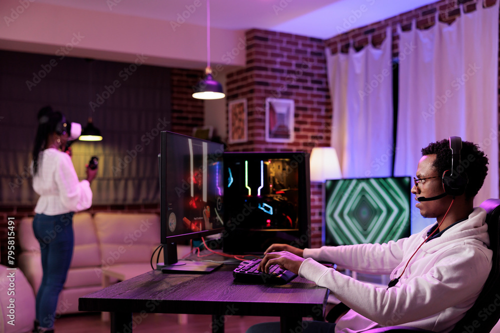 Poster gamer immersed in gaming session at computer desk while girlfriend in background uses virtual realit