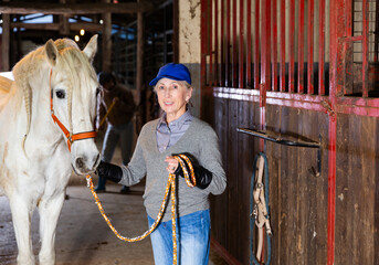 Caucasian senior woman horse breeder standing with white horse in barn and looking at camera.