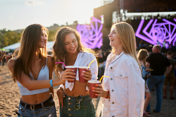 Women clink cups at beach music fest, happy friends toast with drinks in sunshine. Outdoor party vibe, casual summer fun with crowd and festival stage backdrop. Cheerful group celebrate, laughter.