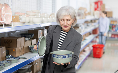 Positive senior female shopper choosing with interest oriental style porcelain soup tureen among variety of dishware arranged on shelves at Asian store