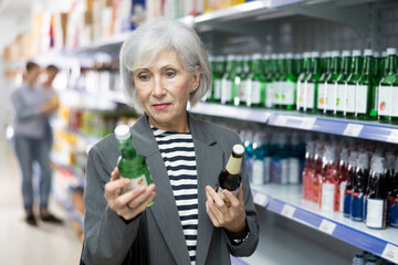Positive senior lady choosing alcoholic beverage at local liquor store, reading label on bottle of...