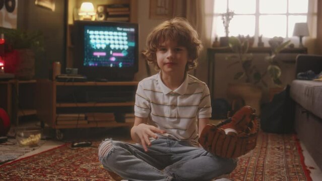 Portrait of happy Caucasian boy sitting on carpeted floor and playing with baseball glove and ball with old space shooter game on tube TV in background at home, 90s