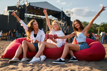 happy girls sit on red bean bag chairs at beach music festival, raise arms, hold drinks. Teen...