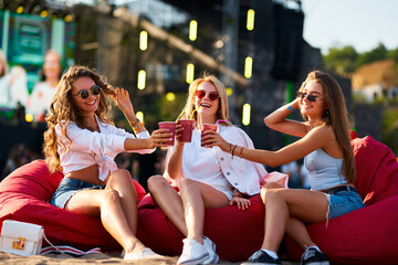 Girls in summer outfits sit on red bean bag chairs, clinking cups at beach music festival. Laugh,...