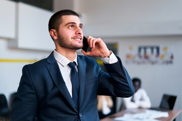 Dressed eastern businessman in suit uses mobile phone in modern office setting. Colleague works at...