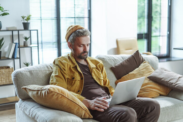 A man in a yellow jacket is sitting on a couch with a laptop in front of him. He is focused on his...