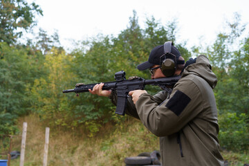 Fototapeta premium A man in a green jacket is holding a rifle and wearing headphones. He is in a forest and he is practicing shooting