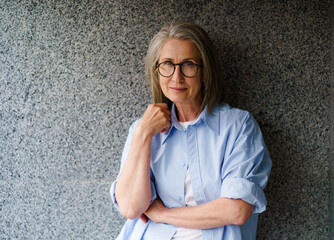 A woman in a blue shirt and glasses is standing in front of a wall. She is looking at the camera...