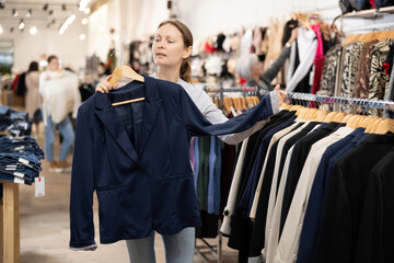 Glad middle-aged female customer holding a hanger with suit jacket in retail outlet - obrazy, fototapety, plakaty