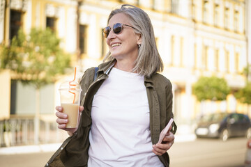 A woman is walking down the street with a cup of coffee and a cell phone in her hand