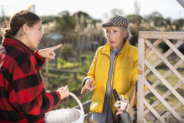 Two upset young and old female neighbors disagree with each other while standing near fence on smallholding in autumn