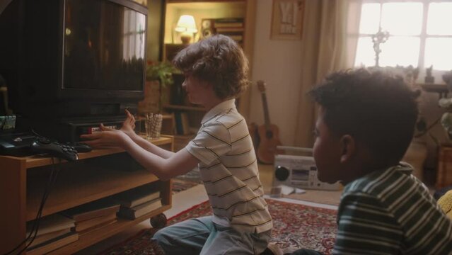 Side footage of Caucasian boy playing cassette with horror movie on tube TV to his African American friend while being alone at home, nostalgic childhood in 90s
