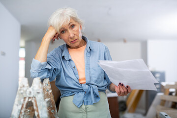 Portrait of frustrated elderly woman interior designer checking documents in apartment during...