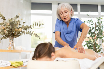 Elderly woman masseuse doing back massage to young woman client in massage room