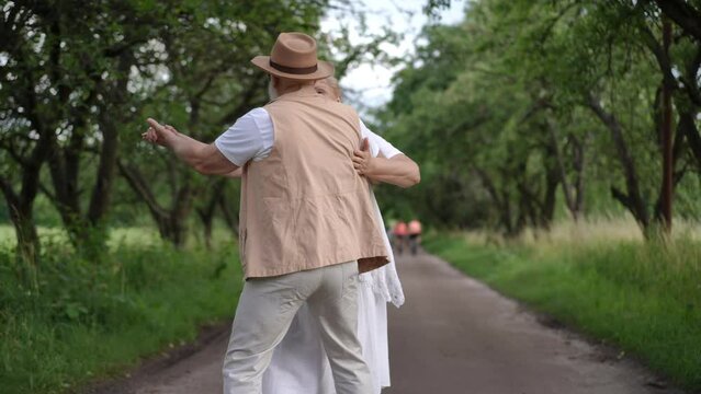 Slow motion. An active seniors having fun spending their leisure, enjoy life and smiling. A happy married couple spins and dances on a park path, holding hands, while walking in a city park on a