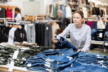 Pleased middle-aged woman choosing jean skirt in clothing store with large assortment