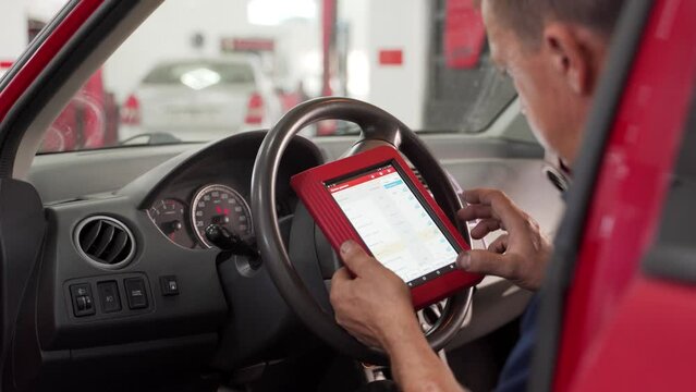 Auto electrician uses OBD2 scanner and tablet to diagnose car in garage. Technician reads fault codes, checks vehicle data. Professional repairman working with diagnostic equipment in workshop.