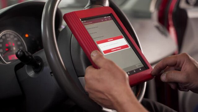 Auto electrician uses OBD2 scanner and tablet for car diagnostics in workshop. Mechanic reads fault codes, performs vehicle maintenance. Technician repairing, checks engine, updates software.