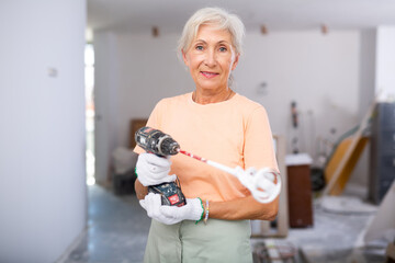 Smiling elderly woman standing in room under reconstruction holding drill with paint mixing...