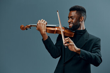 Talented young African American man in stylish black suit playing violin on neutral gray background