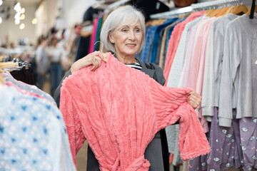 Elderly woman buyer chooses bathrobe in clothing store..
