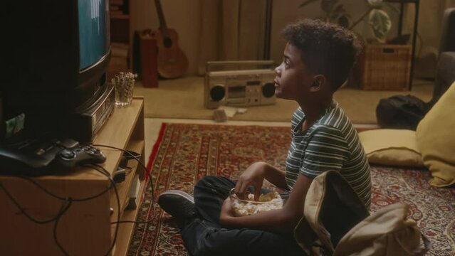 Side handheld footage of gen Y Black boy sitting close to tube TV with bowl of popcorn and absorbedly watching zombie movie being at home alone, carefree childhood in 90s