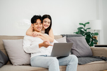 Young Asian couple enjoys a cozy moment on their sofa, interacting with a laptop, in a light-filled, plant-adorned living space. - Powered by Adobe