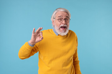 Fototapeta premium Portrait of stylish grandpa with glasses showing ok gesture on light blue background