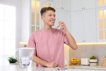 Happy man drinking clear water near filter jug at table in kitchen