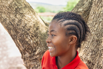 village african teenager girl profile view , wearing braids hairstyle , standing in front of a tree...