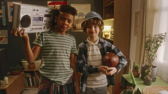 Portrait of two diverse stylish gen Y boys posing with rugby ball and boombox player while standing in cozy retro styled living room and looking at camera