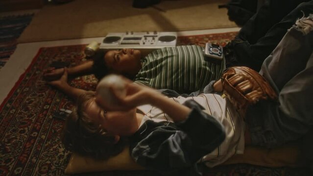 Side footage of 90s little Caucasian boy throwing and catching baseball ball while lying on carpet on floor and chatting with African American best friend at home