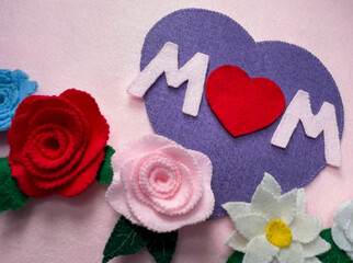 Handmade mother's day card with felt, a purple heart with the word MOM and flowers and roses at the bottom on a pink background. Heart with the word MOM to celebrate Mother's Day. felt texture