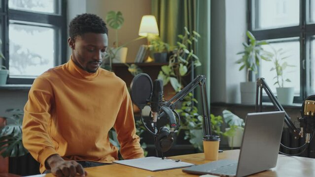 Young African American male blogger putting on headphones, reading notes and speaking into microphone during podcast interview in recording studio. Close-up view, tilt-down shot