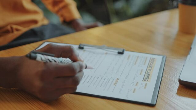 Hands of male podcast presenter proofreading content planner and taking notes on paper at desk in recording studio. Close-up shot