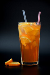 Faceted tall glass of fresh orange juice with ice and straws on a black background with pieces of ripe citrus.