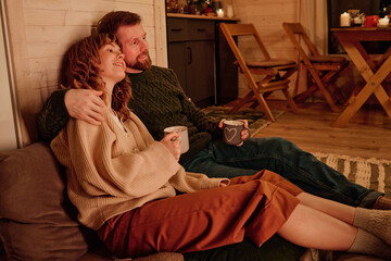 Modern Caucasian man and woman in love holding mugs with tea sitting relaxed on floor in living...