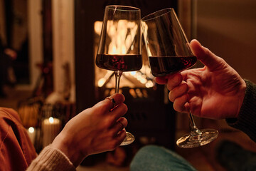 Hands of unrecognizable couple sitting in front of fireplace clinking glasses with wine while having date at home in evening
