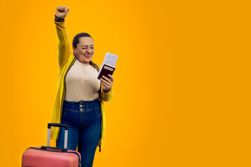 Beautiful woman in summer clothes with pink suitcase, happy with her hand up, celebrating her cheap ticket on yellow background. Tourist very happy to travel abroad with a bargain ticket. Copy space. - obrazy, fototapety, plakaty