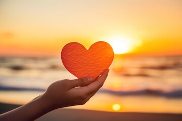 Fototapeta premium Hand holding a cut out of heart shaped paper against a sunset beach sunlight sky tranquility.