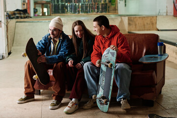 Group of joyful Caucasian boys and girl hanging out together sitting on old couch in skatepark and chatting
