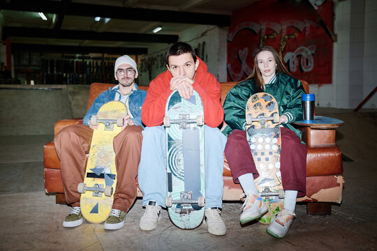 Group of three self-confident gen Z Caucasian skateboard riders in streetwear sitting on couch posing for camera, flash photo